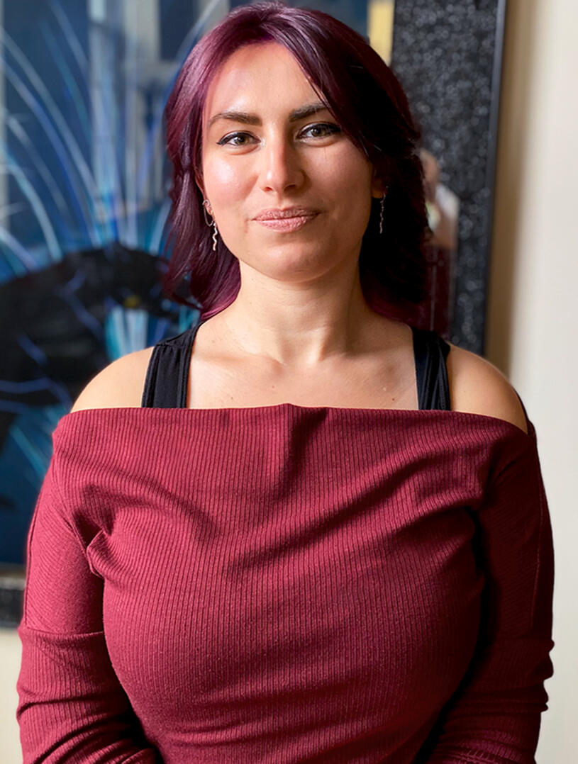 Aleesha Lindstrom - Artist Portrait A vertical portrait of Aleesha Lindstrom, a digital artist, showcasing her unique style. The image features Aleesha in a maroon top standing in front of a framed panther painting.